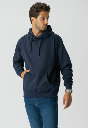 Mand iført marineblå hættetrøje med frontlomme og blå jeans, kigger mod venstre mod en ensfarvet lys baggrund.