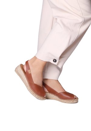 Espadrilles en cuir marron avec bout ouvert, design à bride arrière et semelle en jute tressé ; portées avec un pantalon de couleur claire avec un détail bouton.