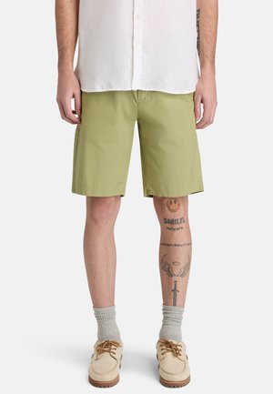 Mężczyzna w jasnozielonych spodenkach do kolan, białej koszuli na guziki, szarych skarpetkach i beżowych butach, pokazujący tatuaże na prawej nodze.