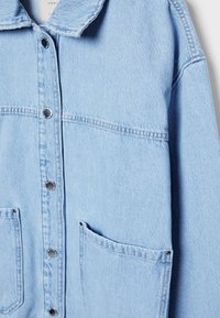 Veste en denim bleu clair avec des boutons-pression argentés, un col et des poches avant couturées sur un fond uni.