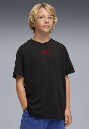 Niño rubio con ojos azules, vestido con una camiseta negra con un pequeño gráfico rojo y pantalones cortos azules, de pie con una mano en el bolsillo.