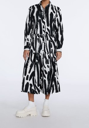 Persoon in een zwart-wit abstract patroon lange mouwen, knoopsluiting midi-jurk met witte sokken en dikke witte loafers.