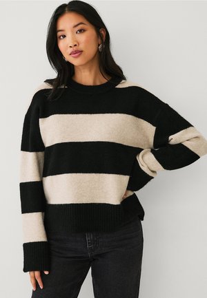 Kvinde iført sort og beige bredstribet sweater med lange ærmer, kombineret med sorte bukser, stående op ad en ensfarvet væg.