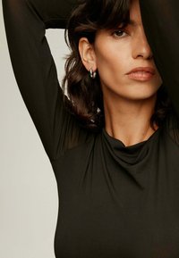 Top negro de manga larga con escote drapeado y acentos de malla transparente. Modelo lleva aretes en forma de aro, con el rostro y los hombros parcialmente visibles.