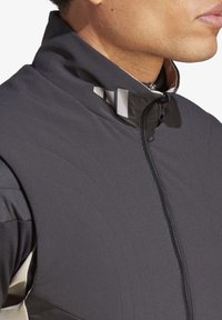 Chaqueta deportiva negra con cuello alto, cierre de cremallera, textura con patrón y acentos reflectantes en el cuello.
