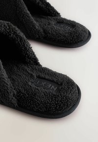 Svarta fluffiga tofflor med mjukt, plyschmaterial. Öppen tå-design med en dämpad sula och en texturerad yta. Logotyp inbäddad i innersulan.