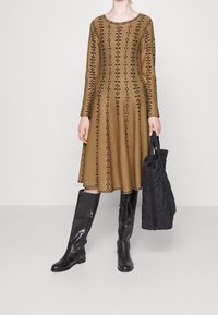 Knee-length, långärmad klänning i tanfärgad stickad tyg med svarta blommönster. Kombinerad med blanka svarta knähöga stövlar och en svart tote-väska.