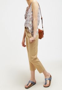 Pantalons beiges coupés, haut sans manches à motifs, sac bandoulière marron et sandales bleues. Les matériaux mettent en avant des tissus légers et un design décontracté.