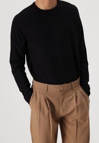 Man med en svart texturerad långärmad skjorta, instoppad i högmidjade ljusbruna veckade byxor, med händerna i fickorna.