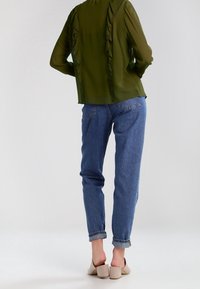 Blusa verde oliva, semi-trasparente, con volant lungo la schiena e le maniche, abbinata a jeans in denim azzurro arrotolati e tacchi beige.