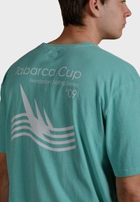 Camiseta de algodón turquesa con un gráfico en blanco. El texto dice "Tabarca Cup Trendplant Sailing Series '09" con acentos de arte lineal.