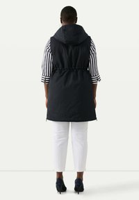 Gilet nero senza maniche con cappuccio, vita affusolata e dettagli con zip, indossato sopra una maglietta a righe orizzontali, abbinato a pantaloni bianchi.