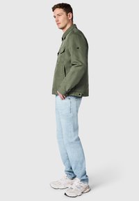 Homme portant une veste vert olive, un jean bleu clair et des baskets blanches, debout en pose latérale décontractée sur un fond uni.