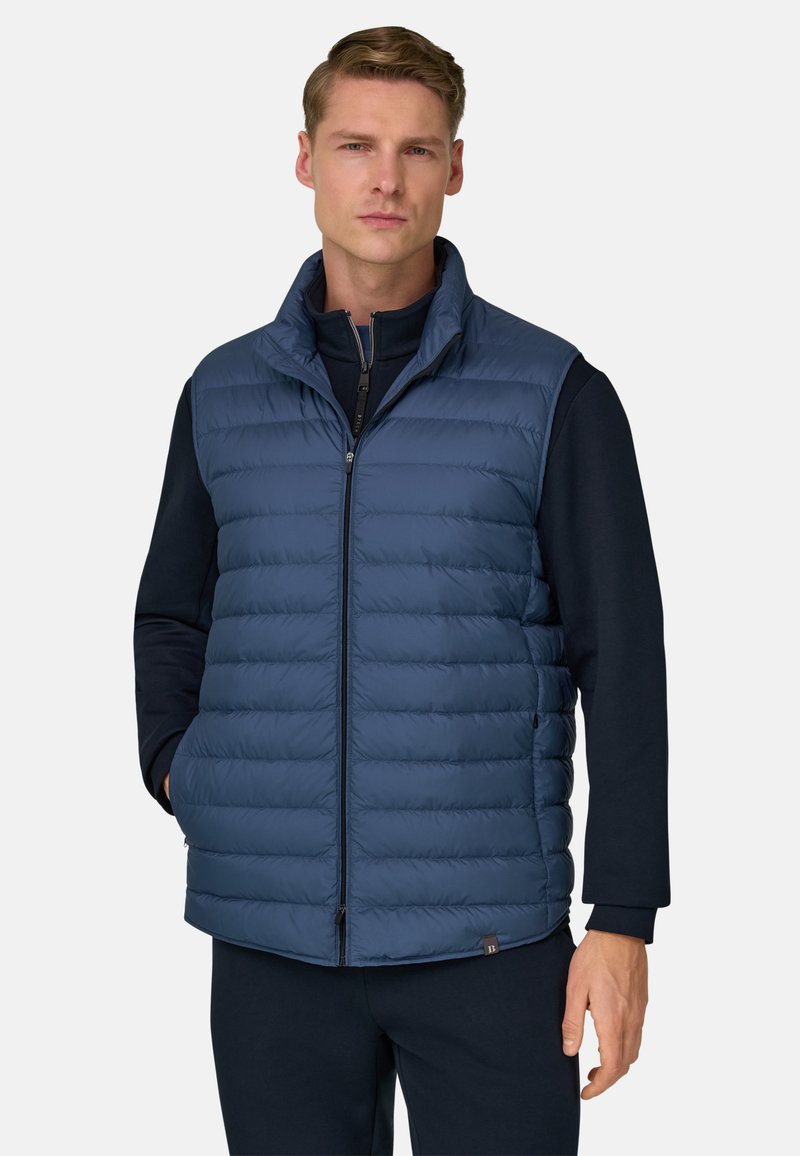 Smanicato imbottito blu navy con collo alto, chiusura a zip, cuciture orizzontali e tasche laterali, indossato sopra una maglia scura.