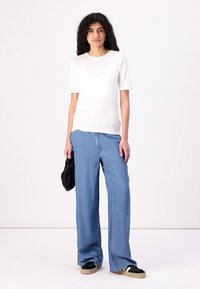 Weißes T-Shirt mit kurzen Ärmeln, kombiniert mit hellblauen weit geschnittenen Jeans. Das Model hält eine schwarze Clutch-Bag und trägt schwarz-weiße Sneakers. Glatte Textur.