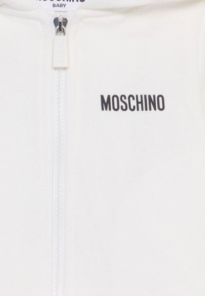 Sudadera blanca con cremallera para bebé, confeccionada en tejido suave. Presenta un logotipo negro "MOSCHINO" en la parte superior izquierda. Textura suave y diseño minimalista.
