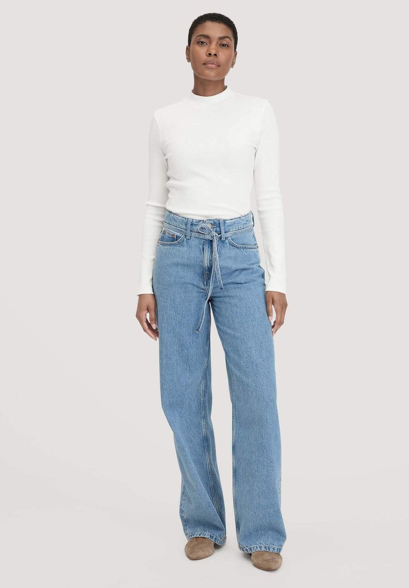 Korkeavyötäröiset vaaleansiniset farkkuhousut, joissa on leveät lahkeet, yhdistettynä valkoiseen pitkähiharaiseen puseroon. Varustettu solmittavalla vyöllä ja hienovaraisella tekstuurilla.