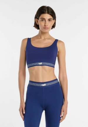Vrouw staand, gekleed in bijpassende blauwe sport-bh en leggings met witte gestreepte banden en New Balance-logo op een witte achtergrond.