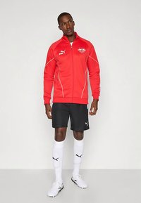 Mees, kes kannab punast Puma kapuutsiga jakki, musti Puma lühikesi pükse, valgeid Puma põlvikuid ja valgeid jalgpallikingi, seisab ühetaolise tausta ees.