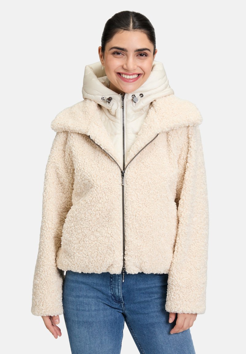Veste en fausse fourrure couleur crème avec un col montant et de larges revers, dotée d'une fermeture éclair argentée et d'une capuche légère et bouffante en dessous.