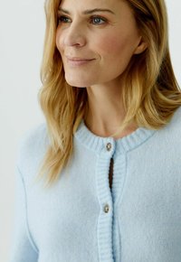 Frau mit blonden Haaren, die eine hellblaue, geknöpfte Strickjacke trägt und leicht zur Seite blickt vor einem einfarbigen Hintergrund.