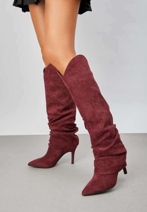 Bottes en suède bordeaux jusqu'aux genoux avec un design flasque, bout pointu et talon aiguille de 4 pouces. Présente un revers plié en haut.