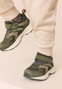 Žalios spalvos sportiniai bateliai iš tinklelio ir zomšos medžiagų, su juodais ir pilkais akcentais, raišteliais ir lipniu dirželiu. Dėvimi su smėlio spalvos laisvalaikio kelnėmis.