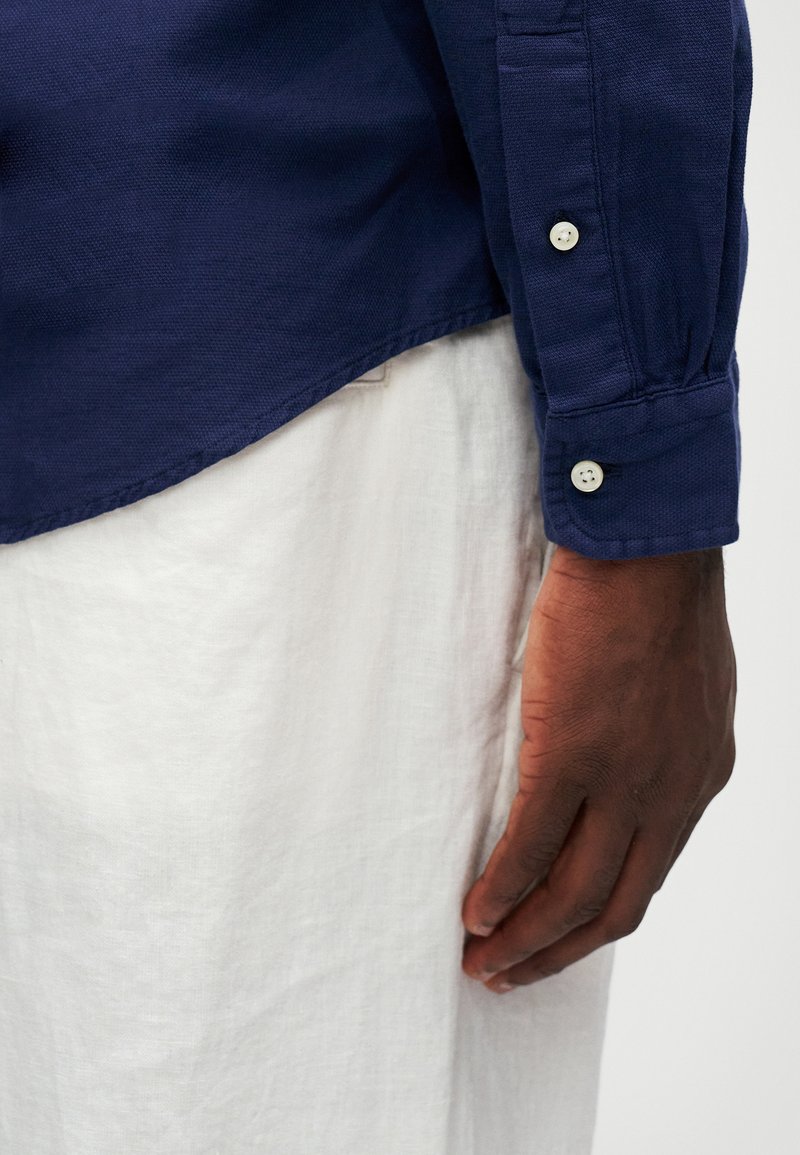 Marinblå langærmet skjorte i struktureret stof med en bølget kant og knapmanchet. Hvide linnedbukser er delvist synlige nedenunder.