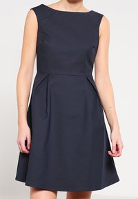 Snygg marinblå klänning med båt-nacke och ärmstycken; utsvängd kjol med horisontella veck vid midjan; slät, enhetlig tyg.