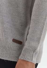 Světle šedý pletený svetr s texturovaným vzorem, pružnými manžetami a hnědou koženou značkovou etikou na dolní straně.