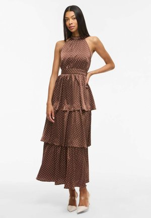 Kobieta o długich ciemnych włosach, ubrana w brązową sukienkę bez rękawów z białymi groszkami i beżowe szpilki w kształcie migdałowego noska, stojąca z ręką na biodrze.
