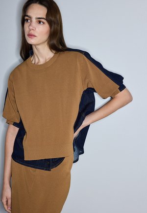 Kobieta o długich brązowych włosach, ubrana w brązowy sweter z dzianiny nałożony na ciemnoniebieską koszulę dżinsową oraz dopasowaną brązową spódnicę, stojąca na tle gładkiej ściany.