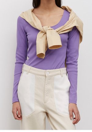 Lila langärmliges Oberteil unter einem beige Stoff, der um die Schultern gebunden ist; cremefarbene Hose mit weißen Seitenstreifen und Fronttaschen.