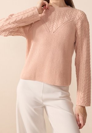 Femme portant un pull en maille torsadée texturée rose clair associé à un pantalon blanc taille haute, posant avec une main près du cou.