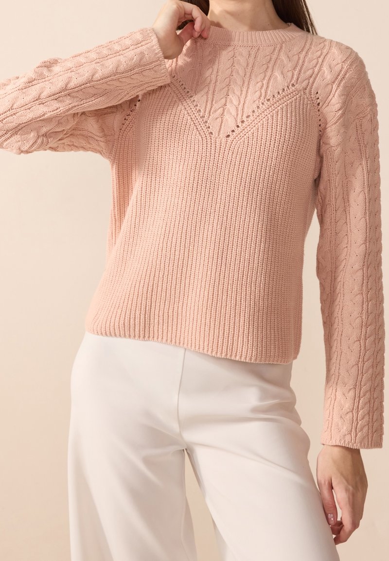 Femme portant un pull en maille torsadée texturée rose clair associé à un pantalon blanc taille haute, posant avec une main près du cou.