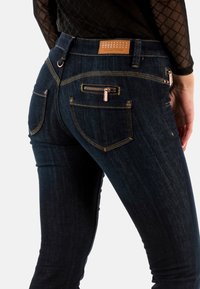 Pantalones vaqueros de mezclilla azul oscuro con un corte ajustado, con costuras doradas, una cintura texturizada y dos bolsillos traseros con detalles de cremallera.
