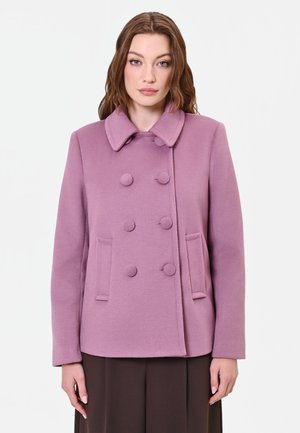 Donna che indossa un cappotto doppiopetto, abbottonato, di colore lilla chiaro con colletto, in piedi davanti a uno sfondo bianco semplice.
