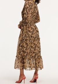 Femme portant une longue robe marron et noire à motifs abstraits, manches longues et volants à étages, associée à des sandales à talons hauts orange.