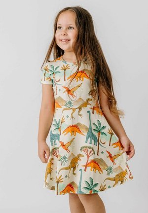 Lächendes Mädchen mit langen braunen Haaren, das ein kurzärmeliges Kleid mit bunten Dinosaurier- und Palmenillustrationen trägt.