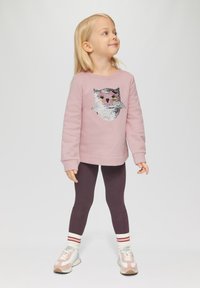 Lichtroze sweatshirt met uilontwerp in pailletten, gecombineerd met donkerpaarse leggings en wit-rode gestreepte sokken. Sneakers met pastelaccenten.