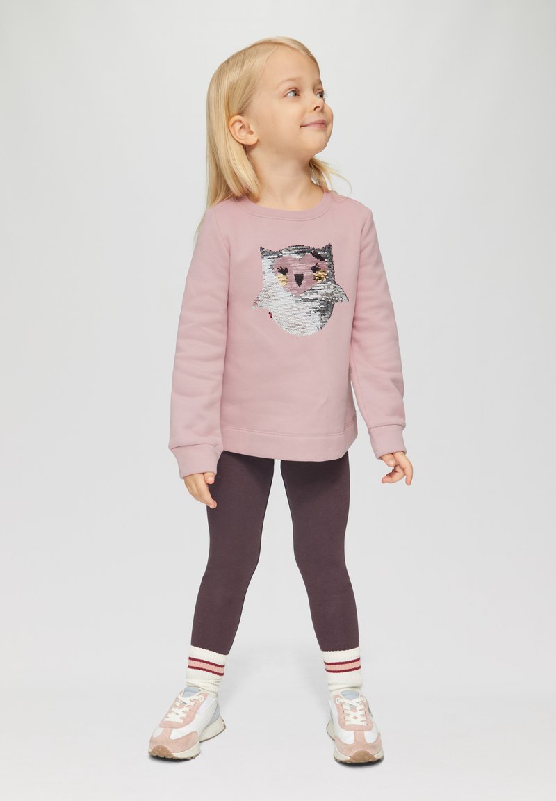 Lichtroze sweatshirt met uilontwerp in pailletten, gecombineerd met donkerpaarse leggings en wit-rode gestreepte sokken. Sneakers met pastelaccenten.