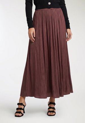 Donna che indossa una lunga gonna marrone plissettata abbinata a sandali neri con tacco alto e tre cinturini e un maglione nero con un bottone decorativo.