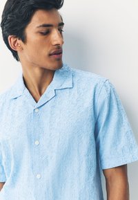 Chemise bleu clair à manches courtes avec un motif floral texturé, une patte de boutonnage à l'avant et un col pointu. Tissu lisse avec des coutures détaillées.