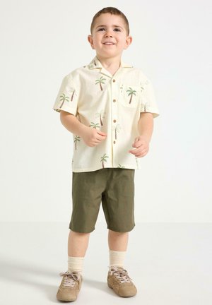 Ung dreng stående iført beige kortærmet skjorte med palmeprint, olivengrønne shorts, cremefarvede sokker og tan sneakers.