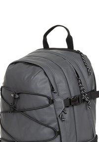 Zaino in nylon grigio con parte superiore arrotondata, lati imbottiti, cordini bungee regolabili e maniglia superiore. Presenta cuciture a contrasto e dettagli neri.
