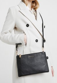 Sort læder crossbody taske med pebble tekstur, guldfarvet hardware og aftagelig strop. Vist mod en hvid frakke.