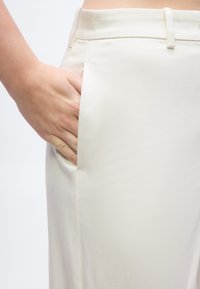 Χέρι που ακουμπάει μέσα στην εμπρόσθια τσέπη ενός ζευγαριού ψηλόμεσων, εκρου, ραμμένων παντελονιών, πάνω σε λευκό φόντο.