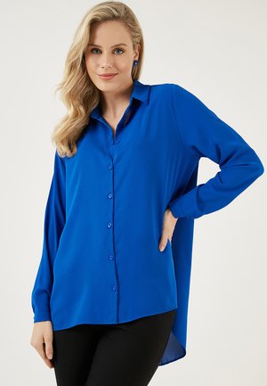 Blue button-up shirt with a collar and long sleeves. Smooth fabric, loose fit, and a slightly longer back hem. Contrast buttons visible.
