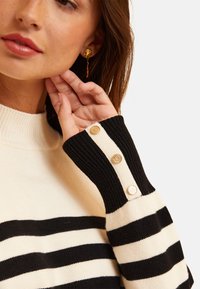 Maglione a righe in crema e nero con maniche a coste, caratterizzato da tre bottoni dorati su una striscia beige. Texture morbida e lavorazione a maglia.