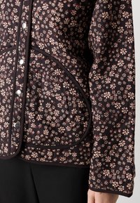 Close-up of dark jacket with small pink floral pattern, black trim, snap buttons, and curved front pocket, worn with black pants.
